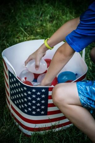 lapsi poimii pienet punaiset, valkoiset ja siniset vesipallot altaasta ja siinä on amerikan lippu