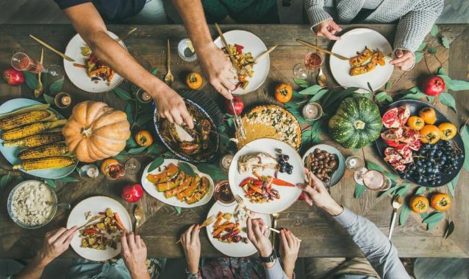 yläpuolella kiitospäiväpöytä, jossa on kalkkunaa, kurpitsapiirakkaa, paahdettuja kauden vihanneksia ja hedelmiä ja ihmisiä täyttämässä lautasia ja syömässä