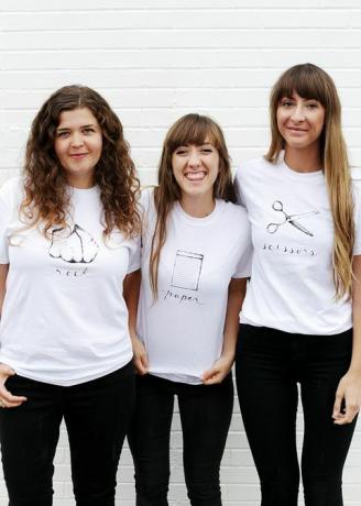 kolme naista valkoisissa t-paidoissa, joissa joko kivi, paperi tai sakset on kirjoitettu ja kuvattu paitaan