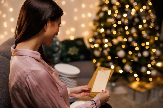 iloinen nuori nainen istuu sohvalla, kimaltelevan joulukuusen vieressä, avaa ja lukee joulukorttia, jonka hän sai lahjansa kanssa