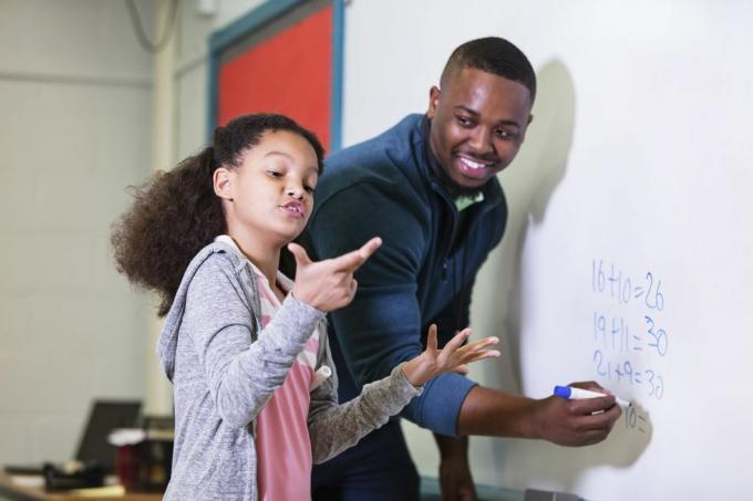 monirotuinen 9-vuotias tyttö matematiikan tunnilla seisoo taululla opettajansa kanssa, nuori afroamerikkalainen mies 20s opettaja katsoo ala-asteen oppilaansa odottaen, kun hän yrittää selvittää vastausta. tyttö