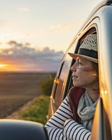 40-vuotias nainen katselee maaseutua matkailuautostaan, hän näyttää tyytyväiseltä ja rentoutuneelta
