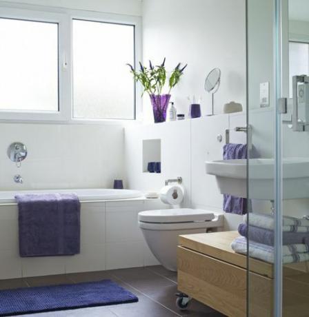Kirkkaasti valaistu kylpyhuone, jossa violetti pyyhe kylpyammeella ja taitetut pyyhkeet lähellä suihkua
