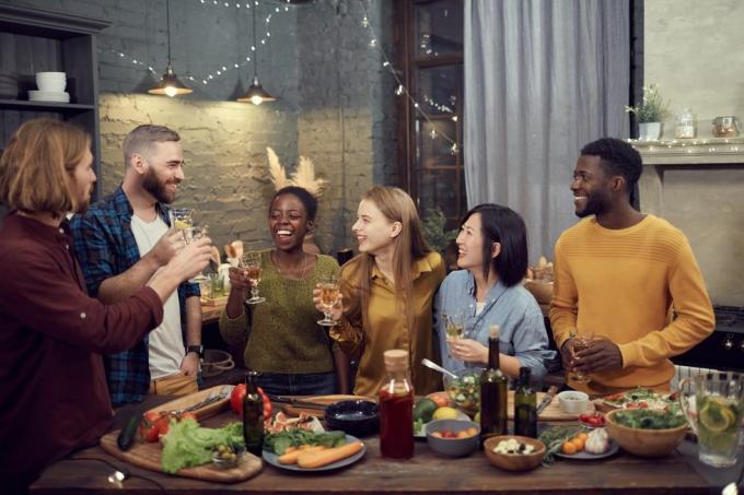 monietninen ryhmä hymyileviä nuoria, jotka nauttivat illallista yhdessä seisoen pöydän ääressä modernissa sisustuksessa ja pitelemässä viinilaseja